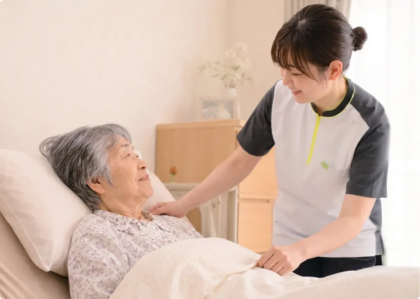 ベッドに横になる高齢女性の肩に女性スタッフが優しく手を添え、微笑みながら声をかけている様子。
