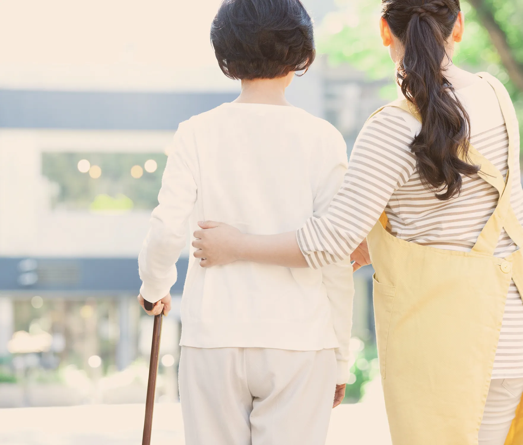 介護士が杖を持つ高齢女性の背中に手を添え、寄り添いながら屋外を歩いている様子。